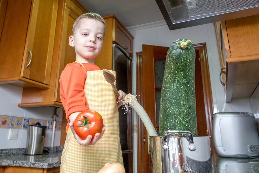 reusing content: shows a young boy in the kitchen with an apron on, getting ready to cook a zucchini that's almost as big as he is. 